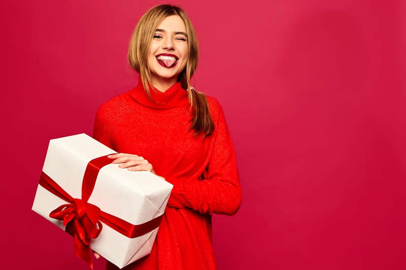 Christmas, x-mas, winter, happiness concept - smiling beautiful stylish woman in sweater. Girl posing on red background. Model with big gift box. Winking and showing her tongue